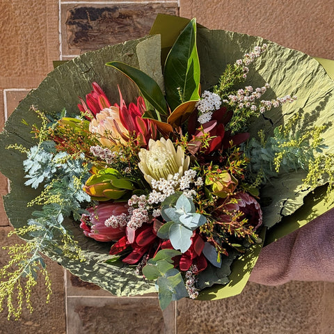 A ceramic vase containing an arrangement of Australian native flowers, including proteas, gum leaves, and possibly a daisy, tied together with a ribbon.